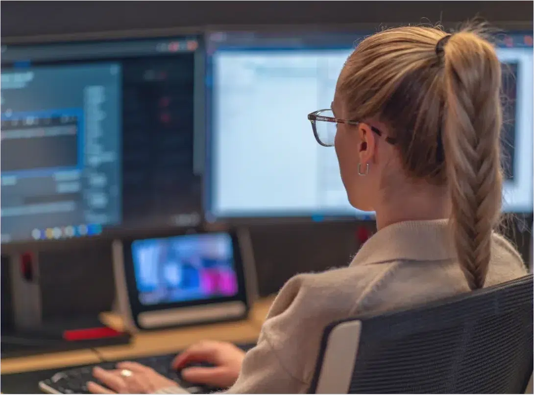 Eine hosttech-Mitarbeiterin mit geflochtenem Pferdeschwanz sitzt an einem Schreibtisch und arbeitet konzentriert an mehreren Monitoren. Auf den Bildschirmen sind Programmier- und Softwareoberflächen für das Server-Monitoring zu sehen. Sie trägt eine Brille und sitzt auf einem ergonomischen Bürostuhl in einem modernen Büroarbeitsplatz