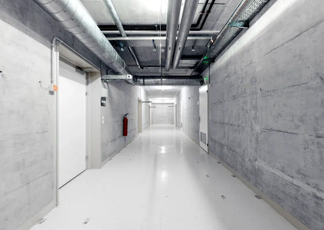 Blick in die unterirdische Bunkeranlage, in welcher das hosttech-Rechenzentrum DATAROCK untergebracht ist. Ein langer Gang mit Beton-Wänden, Lüftungs- und Heizungsrohren an der Decke. Am Ende des Gangs sowie links und rechts in der Wand sind Türen zu sehen. In der Mitte der linken Wand hängt ein Feuerlöscher.