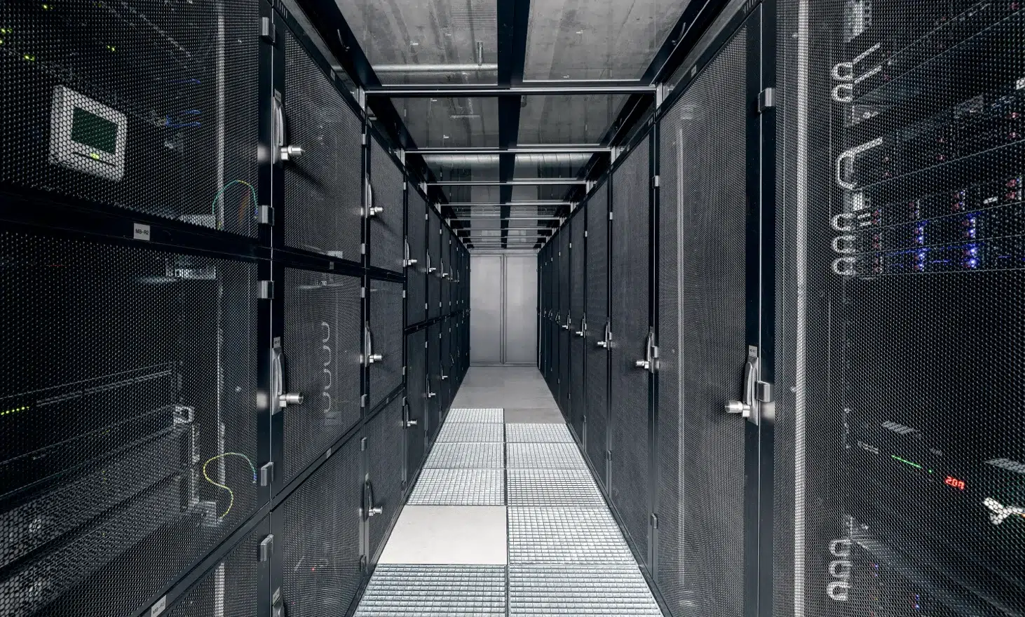 Blick in einen Server-Raum in einem Rechenzentrum von hosttech. Ein langer Gang, bestehend aus Server-Racks, welche mit Gittertüren verschlossene sind. Durch die Gittertüren sieht man auch einige Server. Durch eine Glasdecke ist die Beton-Decke des Raums zu sehen. Der Boden besteht aus Gittern für die Luftzirkulation im Kaltgang.