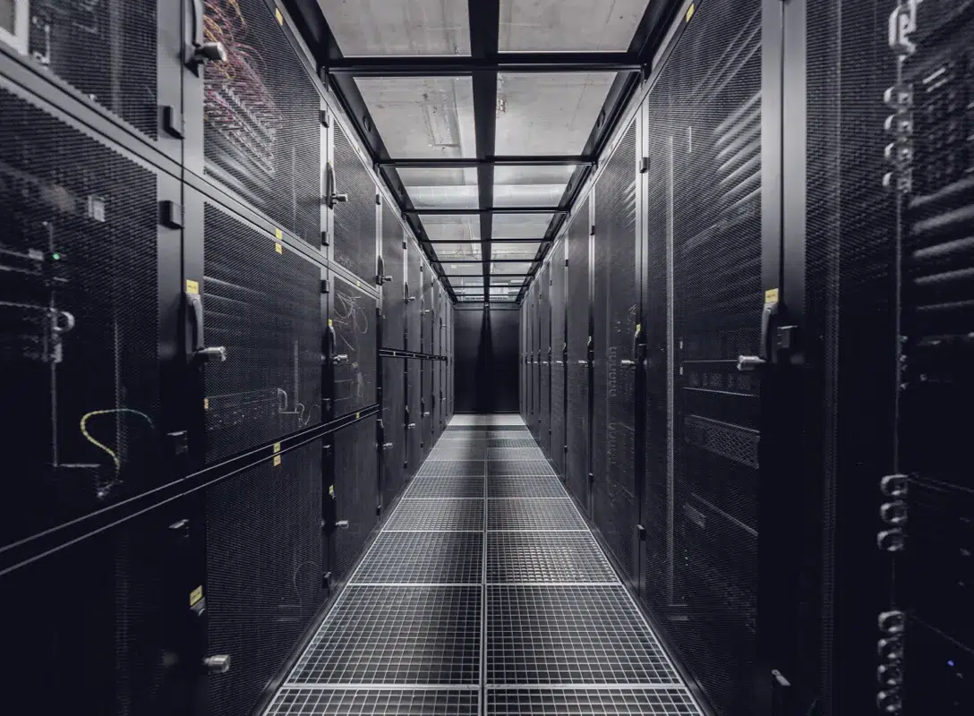 Blick in einen Server-Raum im unterirdischen Rechenzentrum DATAROCK von hosttech. Ein langer Gang, bestehend aus Server-Racks für Colocation, welche mit Gittertüren verschlossen sind. Durch die Gittertüren sieht man auch einige Server. Durch eine Glasdecke ist die Beton-Decke des Raums zu sehen. Der Boden besteht aus Gittern für die Luftzirkulation im Kaltgang.